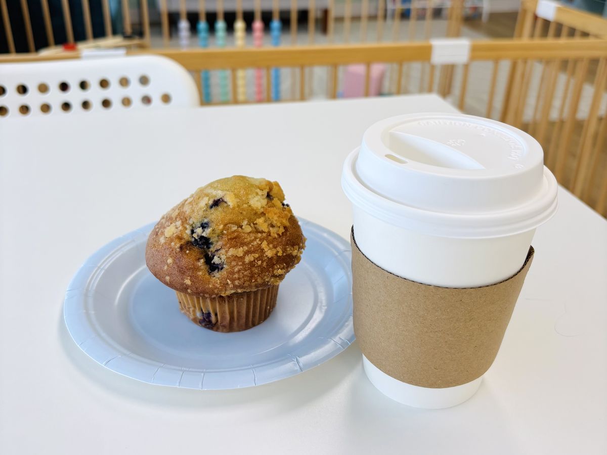A blueberry muffin and a hot coffee on a café table, with the baby area softly blurred behind.