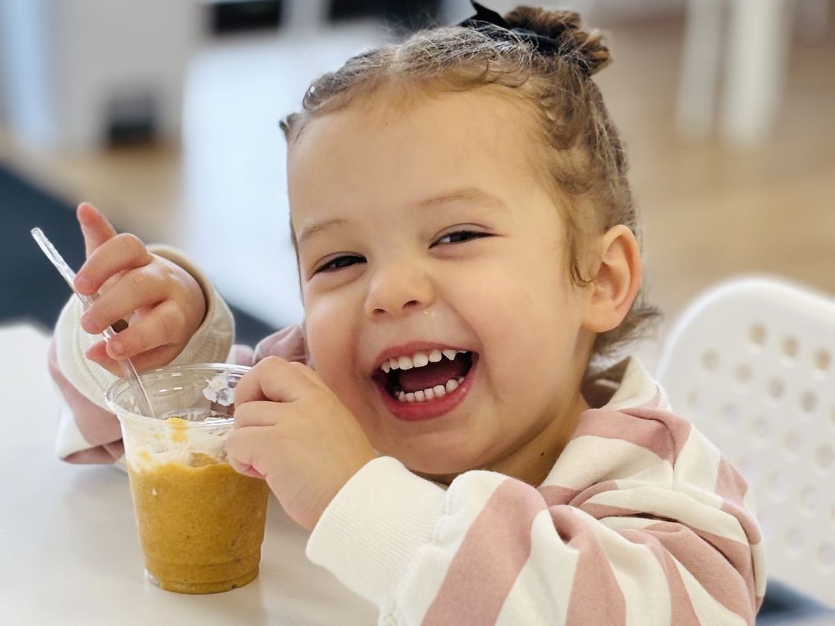 A little girl laughing as she eats a smoothie with a spoon at a café table.