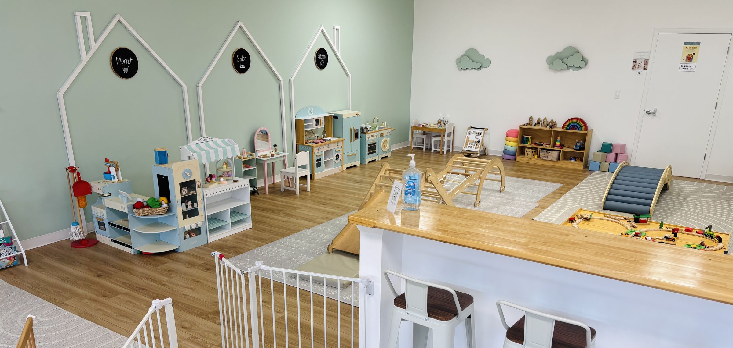 Looking over the café counter and its stools into the play area: pretend-play houses along the mint wall, a climbing arch, a teal roller bridge, a train table and soft-play shapes.