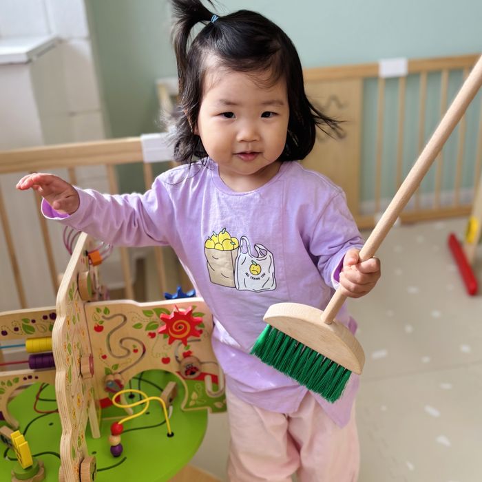 A toddler sweeping with a child-sized broom beside an activity cube.