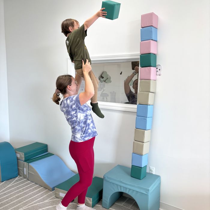 A child lifted up to place the top block on a very tall tower of soft blocks.