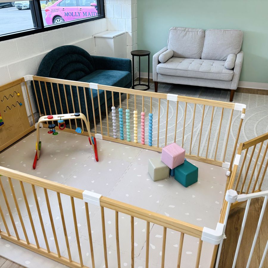 The baby area: a gated play yard on a soft mat with an activity arch, soft blocks and a bead maze, next to a sofa and armchair.