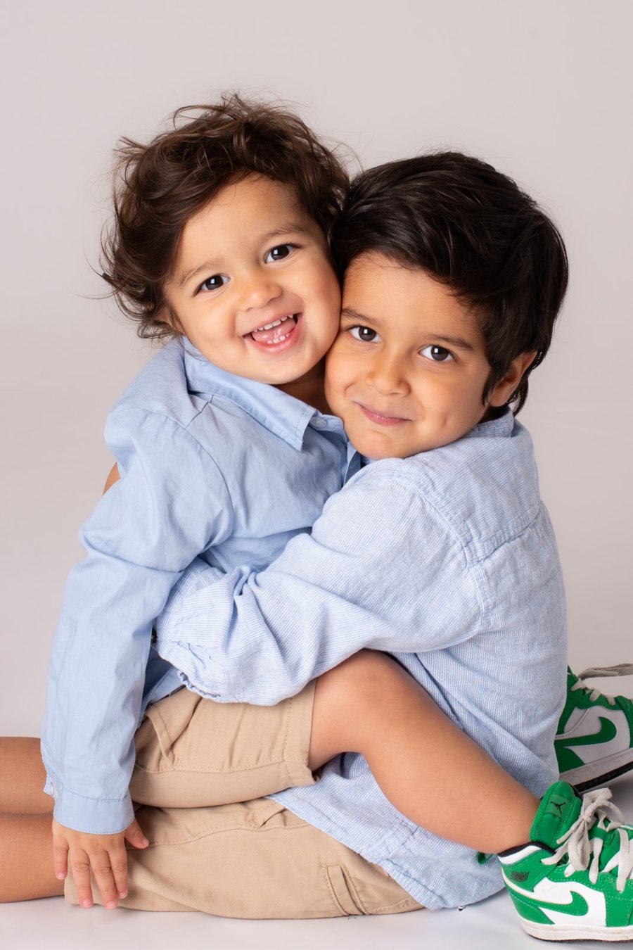 Two young boys in matching blue shirts hugging cheek to cheek and grinning.