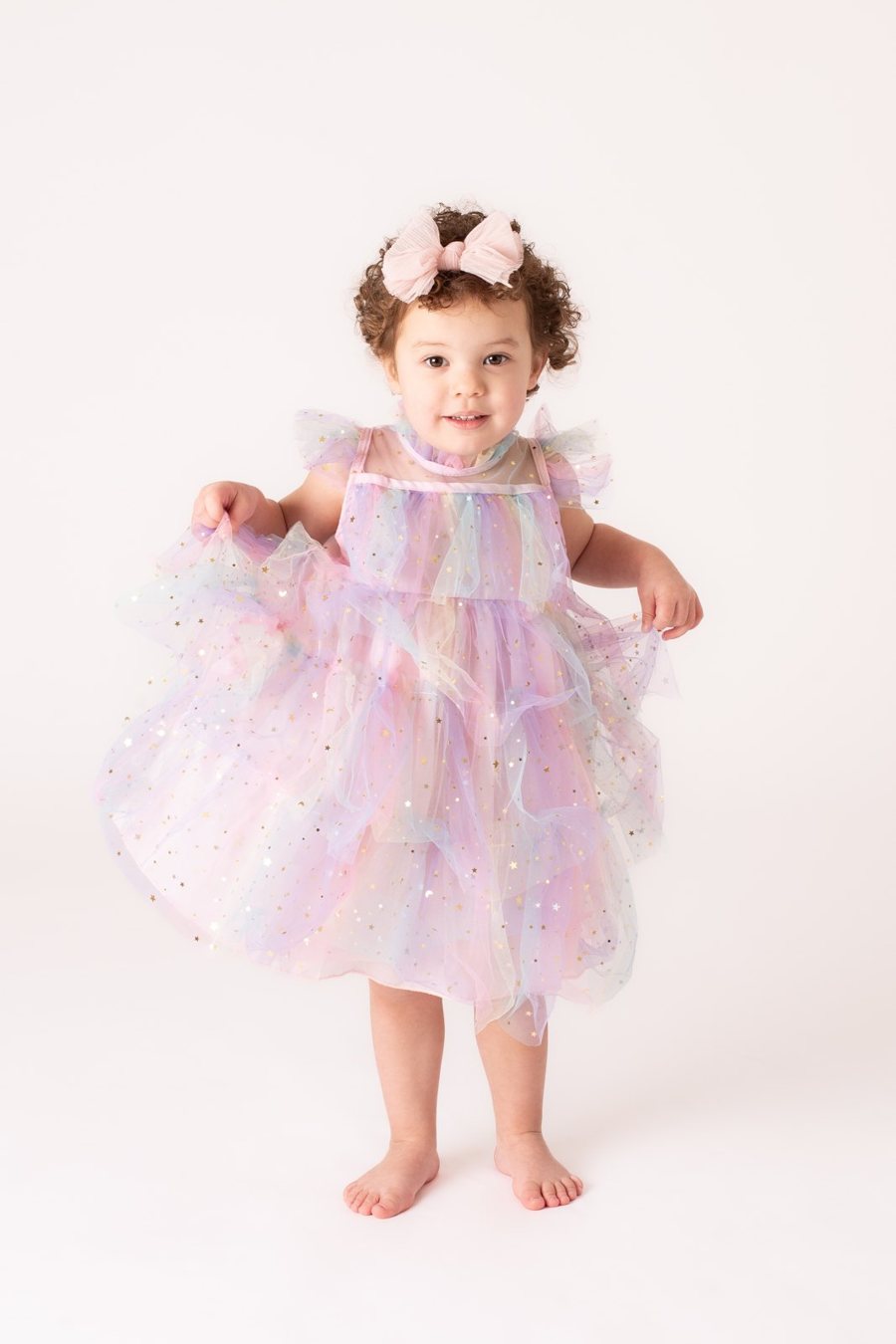 A toddler in a pastel rainbow tulle dress with a flower headband, holding out her skirt in the studio.