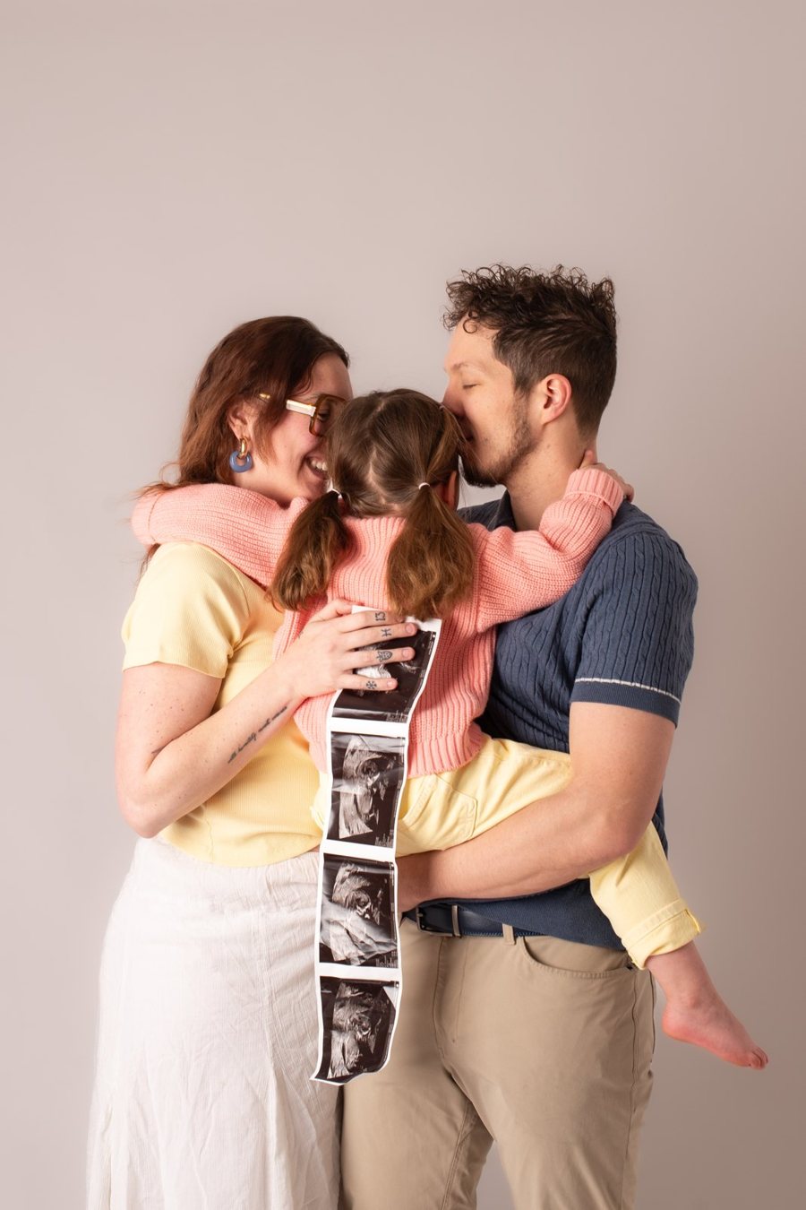Expecting parents hugging their daughter between them, holding a strip of ultrasound pictures.
