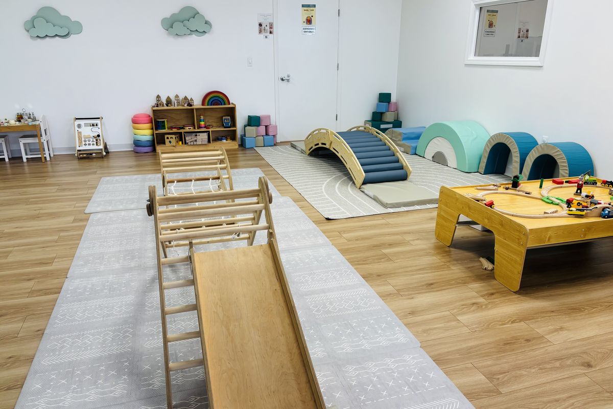 The play floor: a climbing arch and slide, the teal roller bridge, a train table, soft-play rainbows and toy shelves.