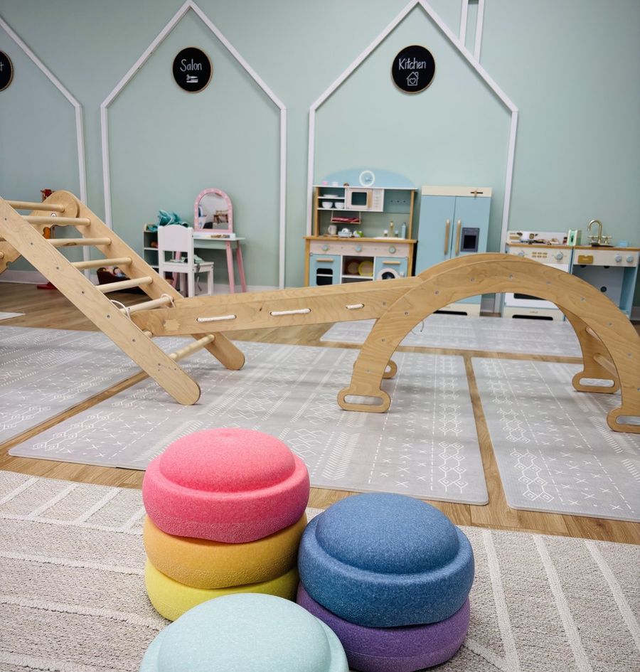 The play floor at Hide and Seek: a wooden climbing arch and ramp on a soft rug, stacking stools in the foreground, and the pretend-play houses along the mint wall behind.