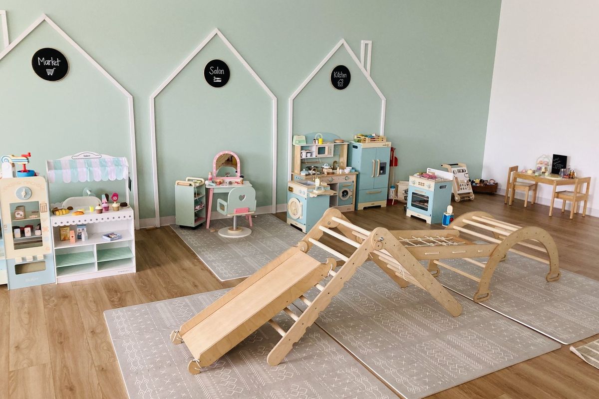 The play floor: a wooden climbing ramp and arch on soft rugs, with a play kitchen, market stall and salon set up under white house frames along a mint wall.
